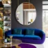 Modern luxury living room by BNG Interiors featuring a curved blue velvet sofa, a large circular wall mirror on a charcoal grey wall, and gold accents with vibrant jewel-tone decor.